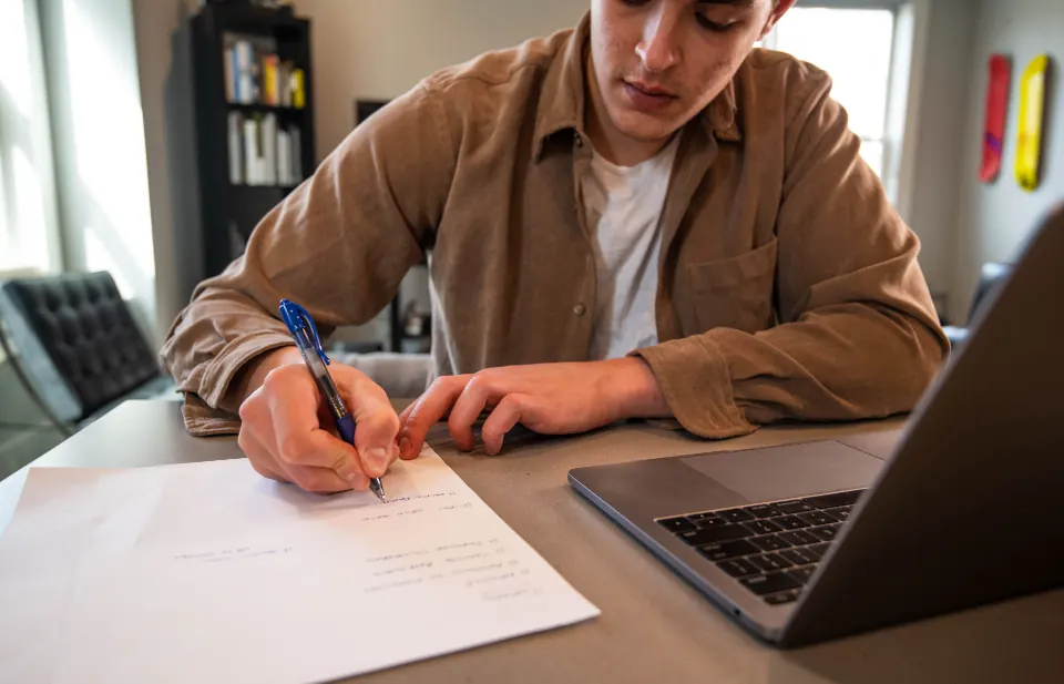 A man working on a laptop and writing notes at a desk