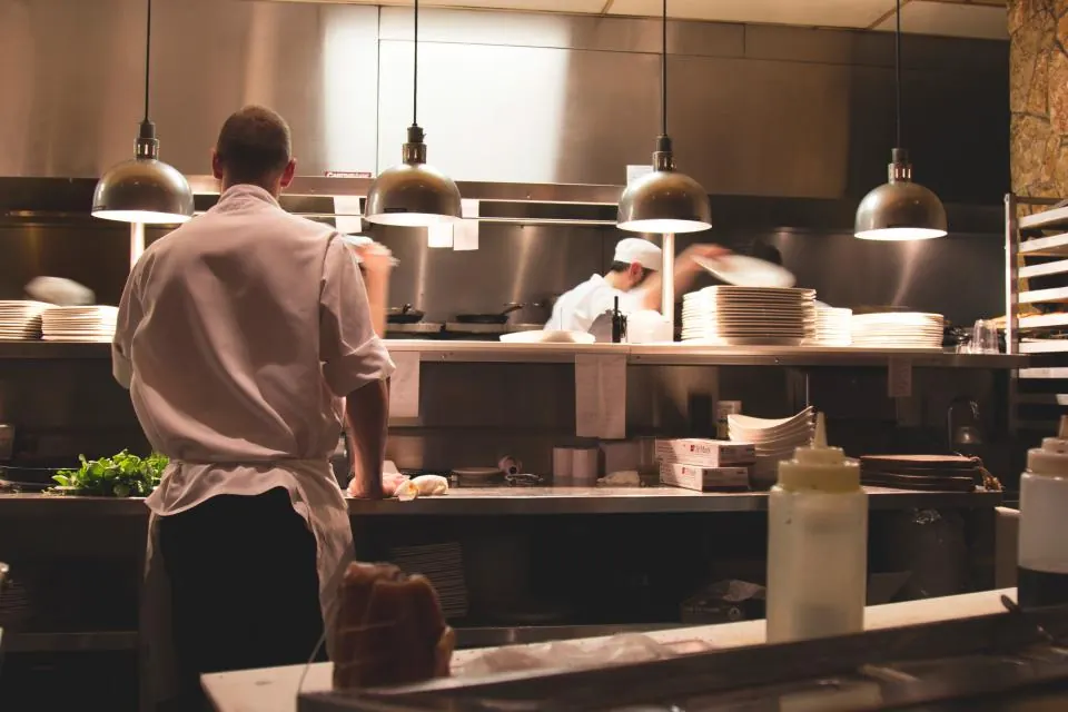 A chef working the pass in a busy restaurant kitchen under pendant lights